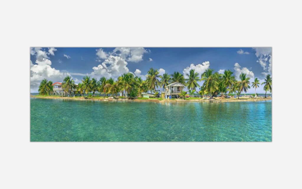 A panoramic image of a peaceful tropical island scene with small houses surrounded by palm trees and calm blue waters under a sunny sky with fluffy clouds.