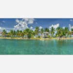 A panoramic image of a peaceful tropical island scene with small houses surrounded by palm trees and calm blue waters under a sunny sky with fluffy clouds.