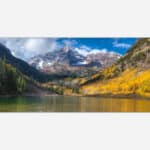 A panoramic photograph of a mountainous landscape with golden yellow aspen trees in autumn and a calm alpine lake in the foreground.
