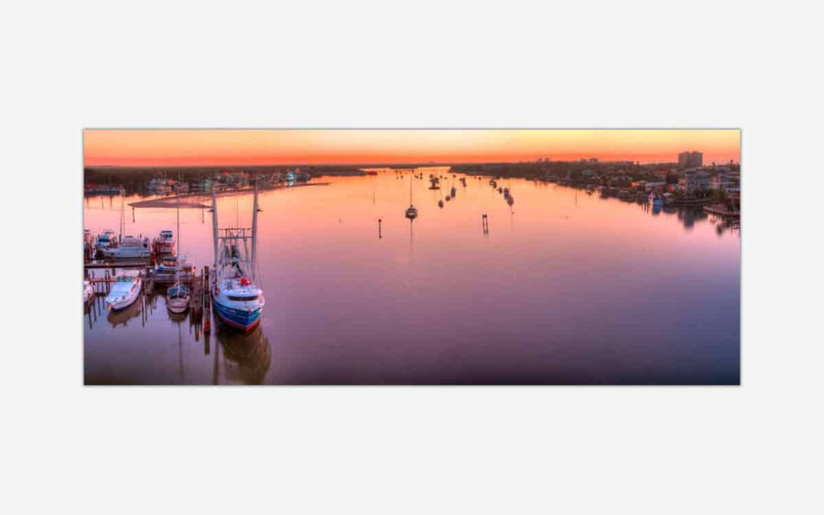 An artwork featuring a peaceful sunset at a marina with multiple boats docked and reflections on calm water, colorful sky in the background, and coastal houses along the shoreline.