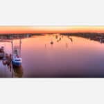 An artwork featuring a peaceful sunset at a marina with multiple boats docked and reflections on calm water, colorful sky in the background, and coastal houses along the shoreline.