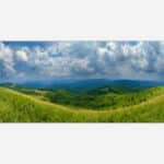 Panoramic landscape photograph of rolling green hills under a cloudy blue sky.