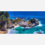 Aerial view of a vibrant coastal landscape featuring a turquoise ocean, white sandy beach, rocky cliffs, and a waterfall amidst lush greenery.