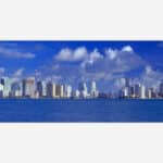 Alt text: An expansive view of a modern city skyline with numerous high-rise buildings along the oceanfront under a blue sky with fluffy clouds.