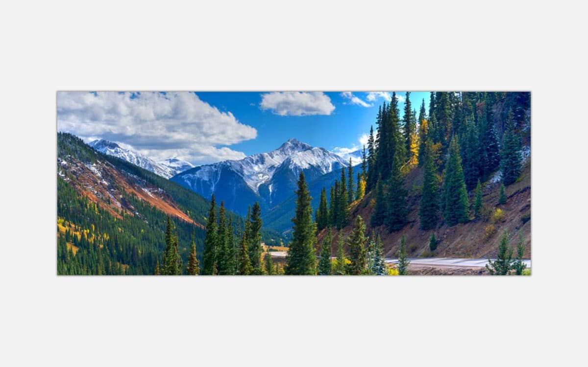 Alt text: A vibrant landscape painting featuring a scenic mountain view with snowy peaks, evergreen pine trees, and autumn colored foliage under a blue sky with clouds.
