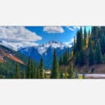 Alt text: A vibrant landscape painting featuring a scenic mountain view with snowy peaks, evergreen pine trees, and autumn colored foliage under a blue sky with clouds.