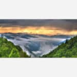 A panoramic landscape photograph of a sunrise over a misty mountain range with lush green forest in the foreground.