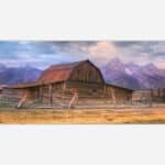 A rustic barn with a wooden fence set against a backdrop of majestic mountains and a warm, colorful sky.