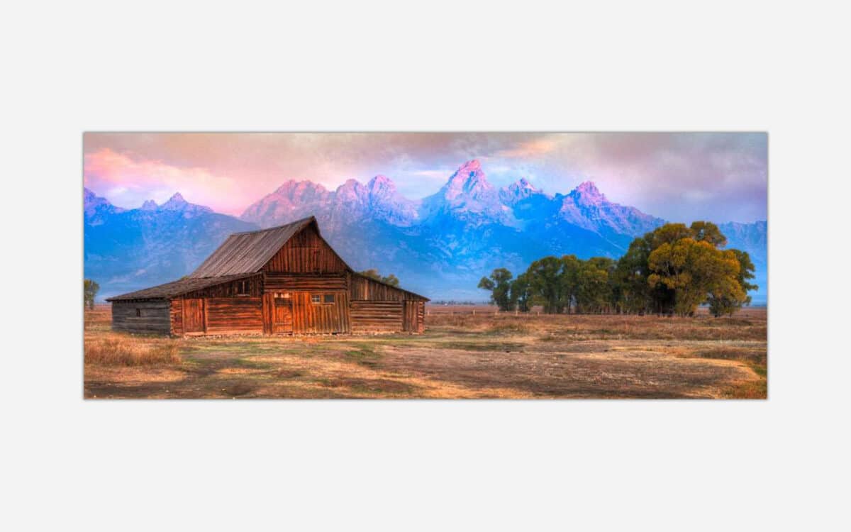 A scenic landscape photograph featuring a rustic wooden cabin with the Grand Tetons mountain range in the background during sunset.