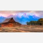 A scenic landscape photograph featuring a rustic wooden cabin with the Grand Tetons mountain range in the background during sunset.