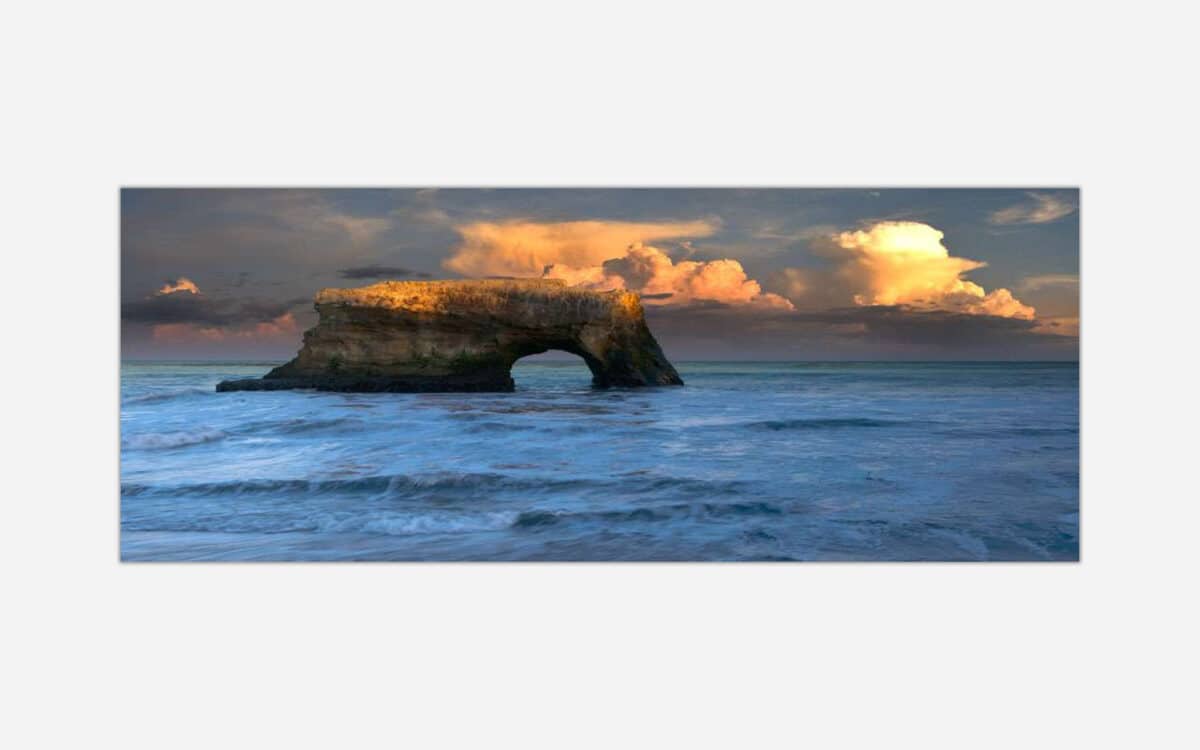 A painting of a natural rock arch in the sea with waves crashing around it as the sun sets, casting warm colors across the clouds and the rock face.