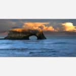 A painting of a natural rock arch in the sea with waves crashing around it as the sun sets, casting warm colors across the clouds and the rock face.