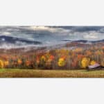 A panoramic photograph of a rustic cabin amidst a colorful autumn forest with misty mountains in the background.