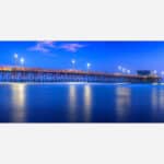 A long exposure photograph of a wooden pier extending into serene blue waters under a twilight sky illuminated by lamp lights.