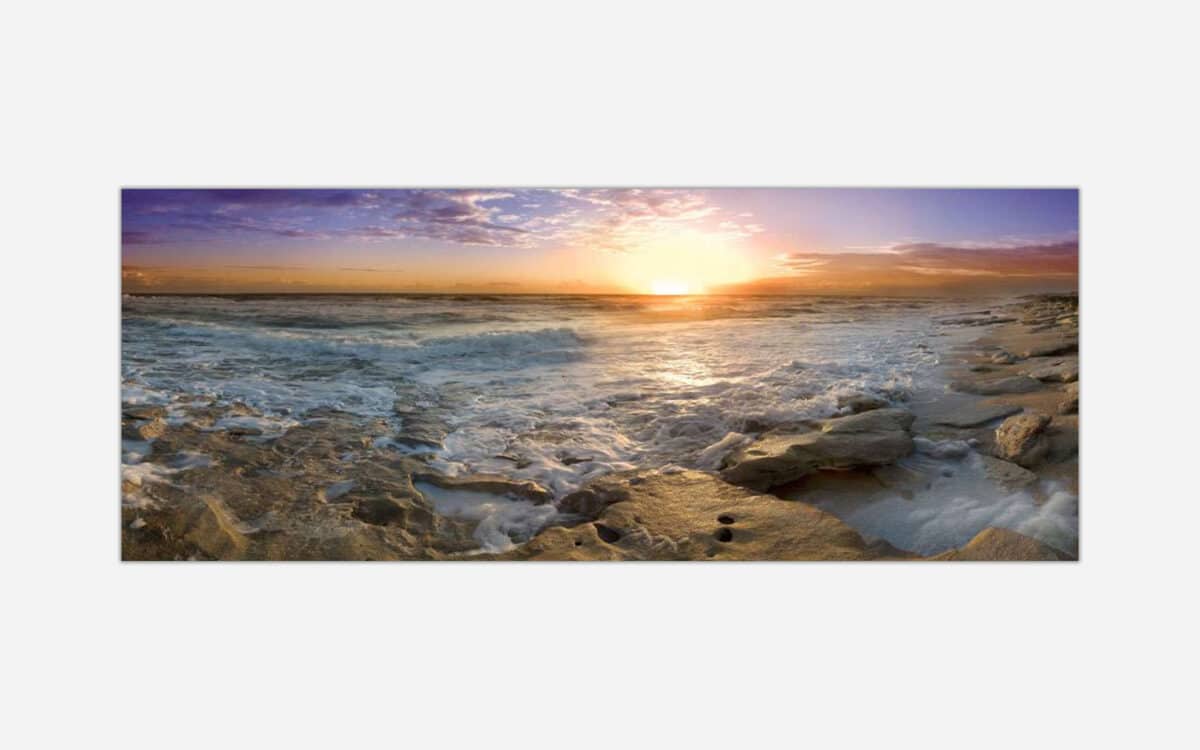 A panoramic seascape photograph of a stunning sunset over the ocean with waves washing over rocky beach foreground.