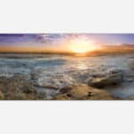 A panoramic seascape photograph of a stunning sunset over the ocean with waves washing over rocky beach foreground.