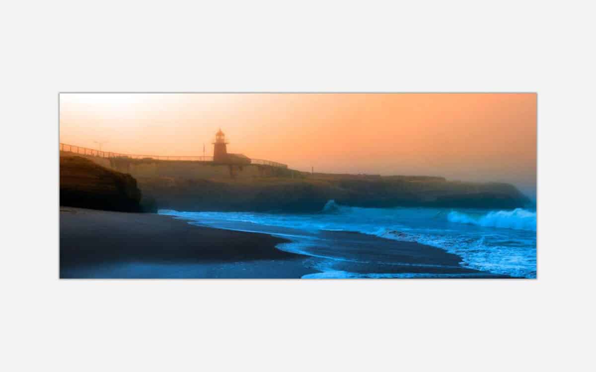 A painting of a sunset view at a coastal scene with a lighthouse overlooking a beach with crashing blue waves.