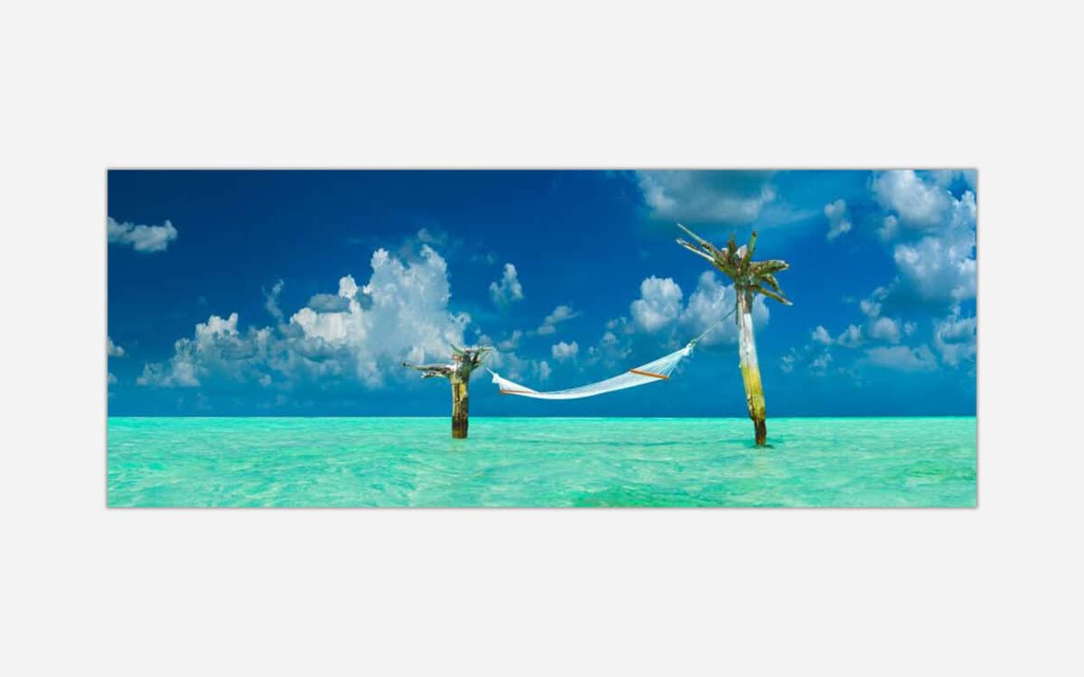 A panoramic wall art of a hammock tied between two palm trees over crystal clear turquoise water under a blue sky with fluffy clouds.