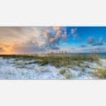 A panoramic view of a beach at sunset with dune grass in the foreground and a colorful sky above the horizon.