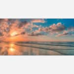 A scenic view of a beach at sunset with a vibrant sky reflected on the ocean and wet sand.