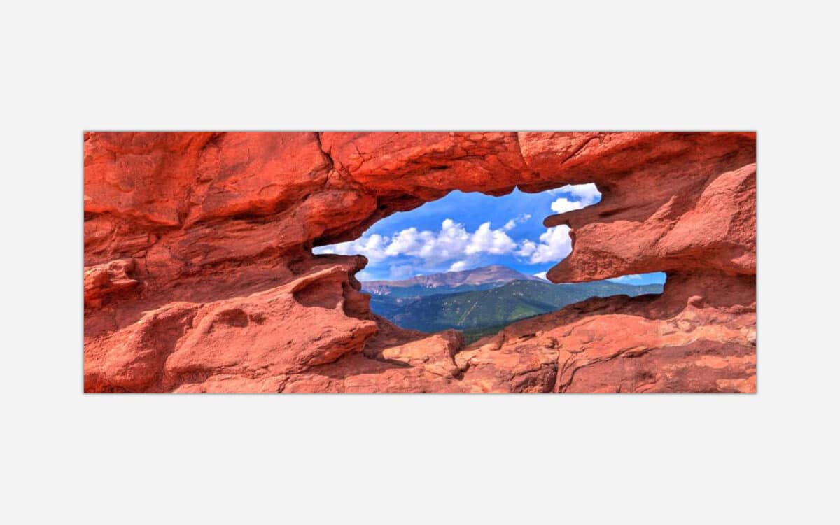 A photograph of a scenic view through a natural red rock arch formation showing a panoramic landscape of mountains and blue sky.