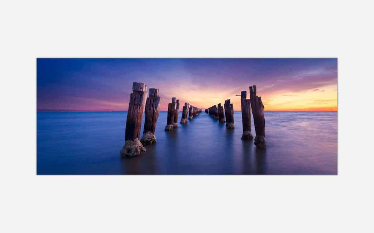A fine art photograph featuring a long exposure of a dilapidated pier stretching into the sea during sunset with vibrant colors in the sky and smooth water surface.