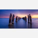 A fine art photograph featuring a long exposure of a dilapidated pier stretching into the sea during sunset with vibrant colors in the sky and smooth water surface.