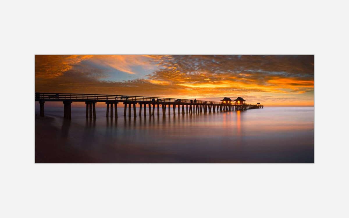 A photograph of a long pier extending into the ocean with a dramatic orange sunset sky and smooth water surface due to long exposure.