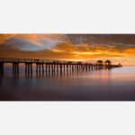 A photograph of a long pier extending into the ocean with a dramatic orange sunset sky and smooth water surface due to long exposure.