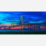 A panoramic photograph of a suspension bridge at dusk with a vibrant and colorful sky in the background.