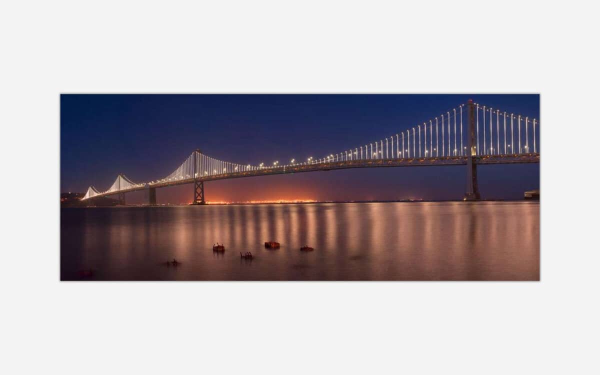 A panoramic photograph of the Bay Bridge in San Francisco with illuminated lights at dusk, reflecting on the water.