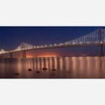 A panoramic photograph of the Bay Bridge in San Francisco with illuminated lights at dusk, reflecting on the water.