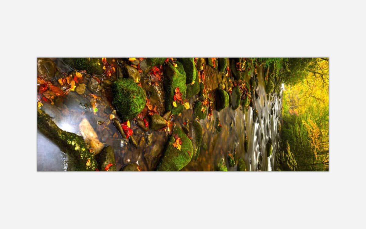 A panoramic photograph of a serene waterfall surrounded by moss-covered rocks and autumn leaves in a forest.