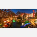 A vibrant night photograph of a city riverwalk area with illuminated bridges, colorful umbrellas, and lively outdoor restaurants.