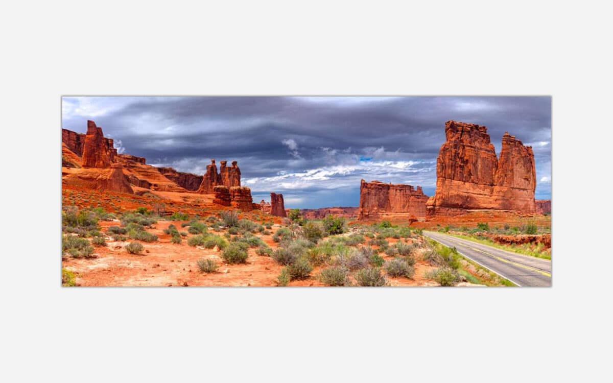 A panoramic photograph of a desert landscape featuring Arches National Park with sandstone formations under a dramatic cloudy sky, a visible road adds depth to the scenery.