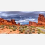 A panoramic photograph of a desert landscape featuring Arches National Park with sandstone formations under a dramatic cloudy sky, a visible road adds depth to the scenery.
