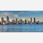 A panoramic photograph of a modern city skyline with skyscrapers and a sailboat on the water at sunset.