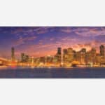 A panoramic cityscape art piece depicting a vibrant sunset over a city with skyscrapers, lit buildings, and a bridge over water.
