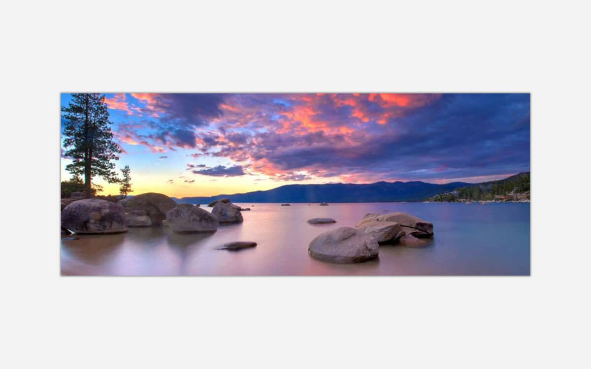 A panoramic photo of a serene lake with colorful sunset skies, silhouetted mountains in the background, and smooth water with rocks in the foreground.