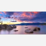 A panoramic photo of a serene lake with colorful sunset skies, silhouetted mountains in the background, and smooth water with rocks in the foreground.