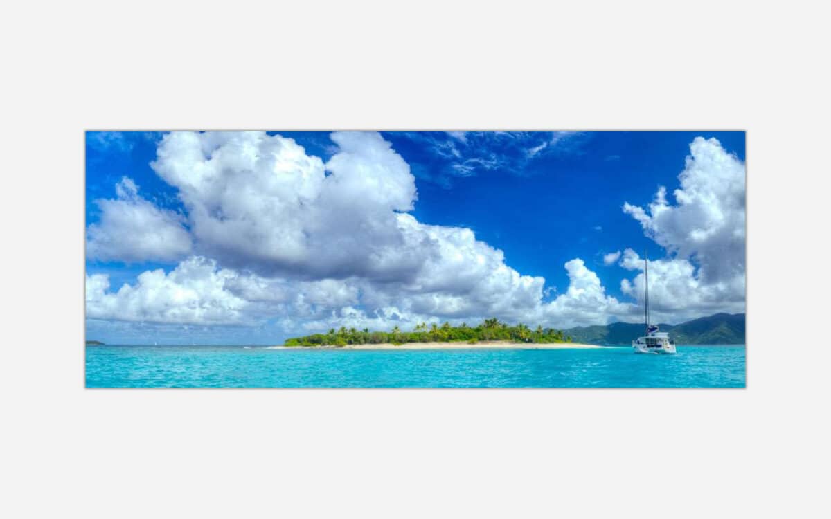 A panoramic view of a serene tropical island with lush greenery and a white sailboat on the clear blue water under a blue sky with fluffy clouds.