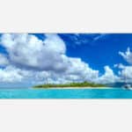 A panoramic view of a serene tropical island with lush greenery and a white sailboat on the clear blue water under a blue sky with fluffy clouds.