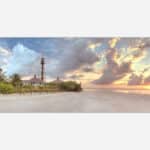 Panoramic view of a tranquil beach scene at sunset with a lighthouse and lush greenery.