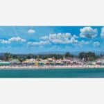 A vibrant art print featuring a seaside amusement park with a roller coaster, colorful buildings, beachgoers, and a clear blue sky.