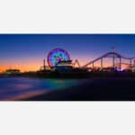 A photograph of a beach pier amusement park at sunset with a colorful Ferris wheel and roller coaster silhouettes against a twilight sky.