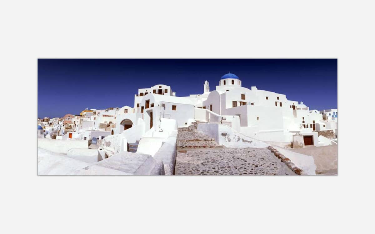 A panoramic photograph of the white painted architecture with blue domes in the village of Oia on the island of Santorini, Greece under a clear blue sky.