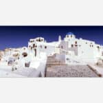 A panoramic photograph of the white painted architecture with blue domes in the village of Oia on the island of Santorini, Greece under a clear blue sky.