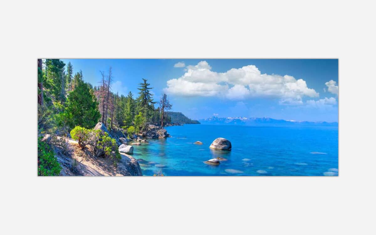 A panoramic landscape photograph of a serene lake view with clear blue waters, pine trees, and distant mountains under a clear sky.