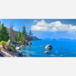 A panoramic landscape photograph of a serene lake view with clear blue waters, pine trees, and distant mountains under a clear sky.