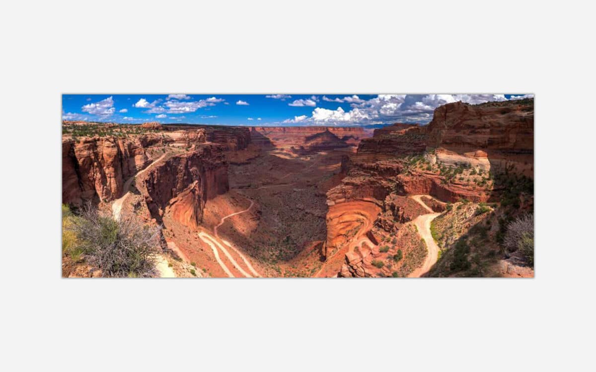 Panoramic photo of a meandering canyon with red rock formations under a blue sky with clouds.